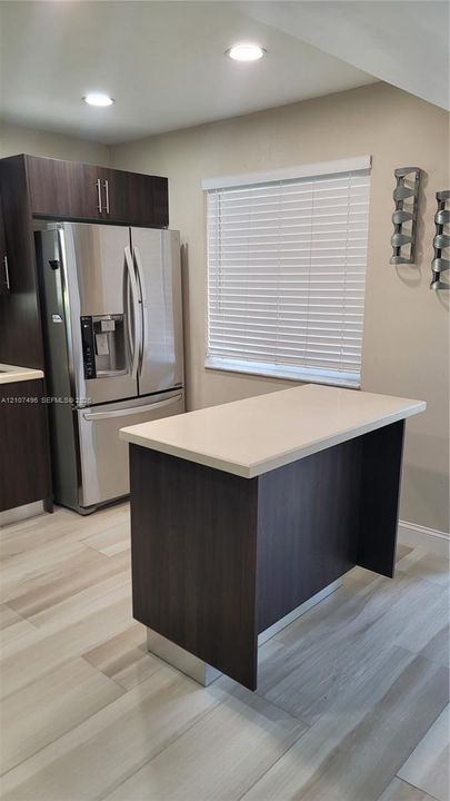 Kitchen featuring dark wood-finish cabinetry, a central island with a stone-composite countertop, stainless steel refrigerator, recessed lighting, and wood-finish tile flooring