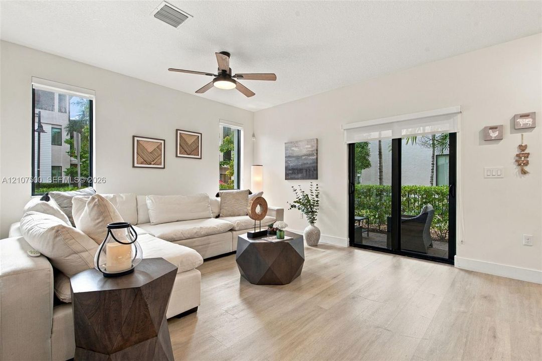 Neutral-toned living space featuring light-colored wood-finish flooring, a ceiling fan with integrated lighting, dual vertical windows, and a sliding glass door providing external access