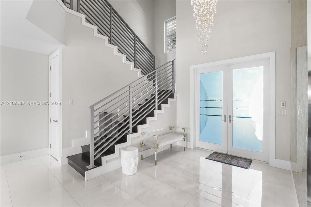 Foyer featuring high ceilings, large-format tile flooring, a double-door entry with frosted glass detailing, a contemporary tiered crystal chandelier, and an open-riser staircase with horizontal metal railing
