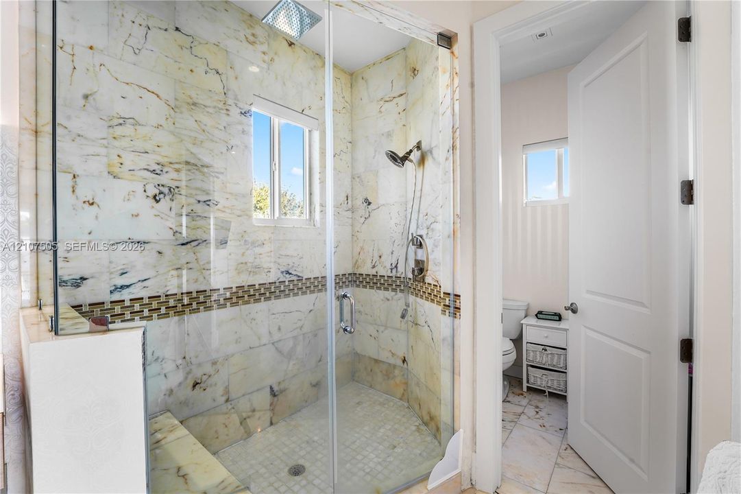 Walk-in shower featuring floor-to-ceiling stone tile surrounds, decorative mosaic tile inlay, built-in stone bench, glass enclosure, and a ceiling-mounted rainfall showerhead