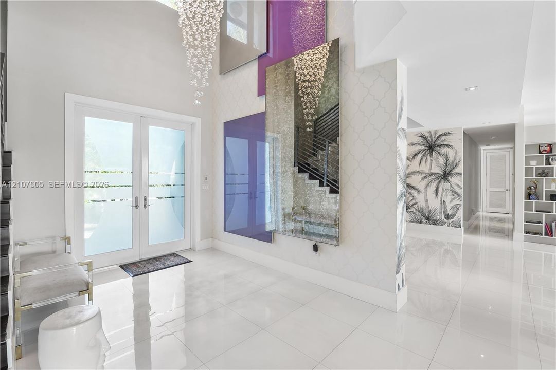 Double-height entryway featuring large-format polished tile flooring, double glass-paneled doors with horizontal frosted accents, a grand tiered crystal chandelier, and custom floor-to-ceiling mirror and glass wall installations