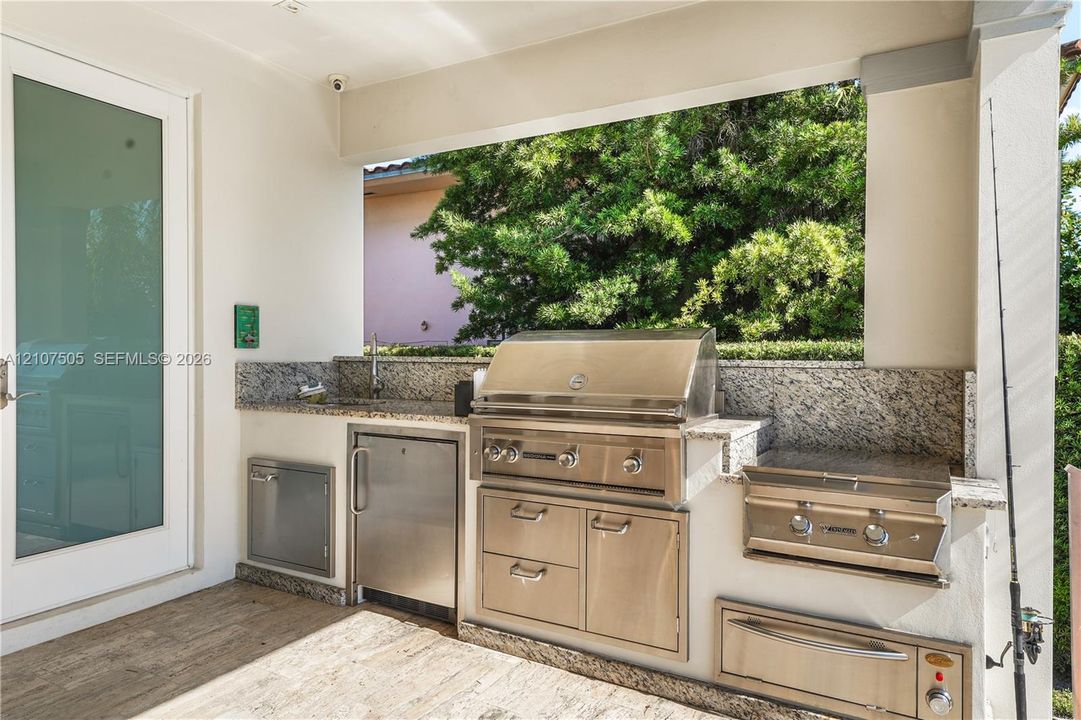 Covered outdoor kitchen featuring stone countertops, built-in stainless steel grill, side burner, under-counter refrigeration, and integrated storage drawers