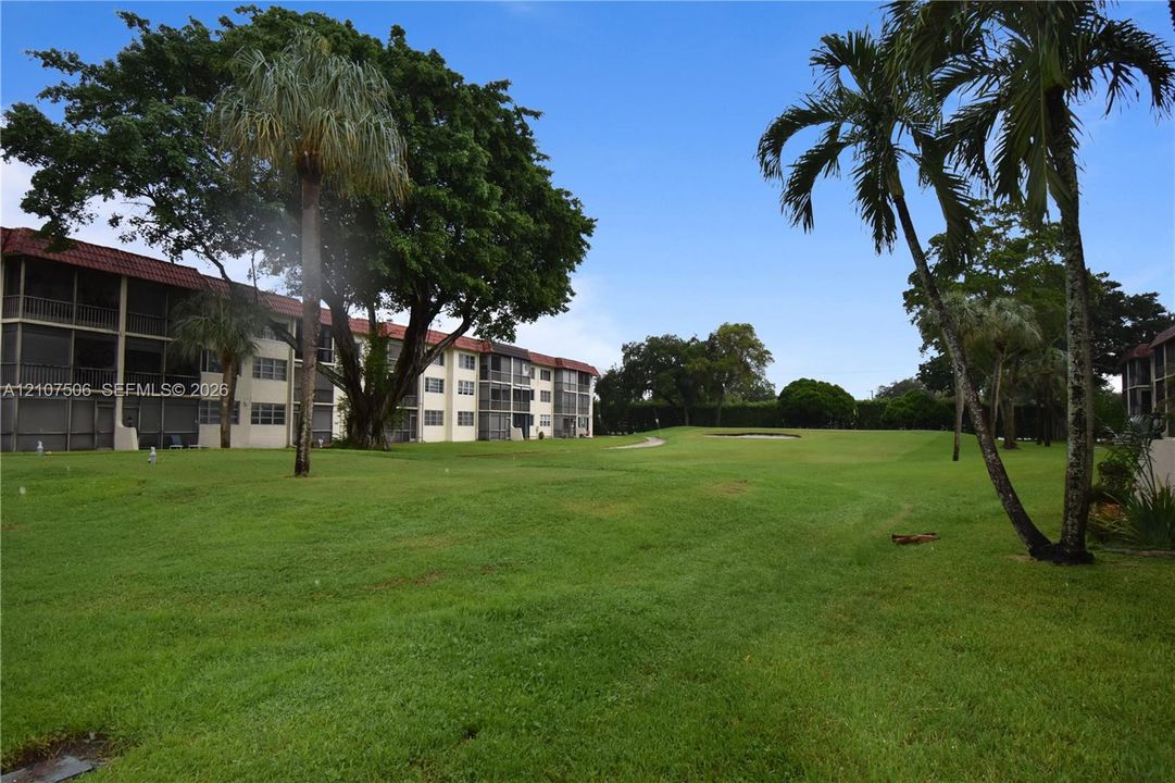 Exterior of a multi-story building featuring screened balconies, expansive green space, mature canopy trees, and established palm trees
