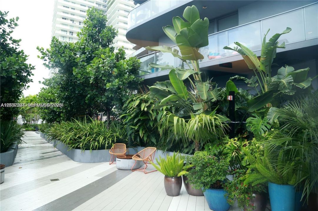Private terrace featuring large-format tile flooring, perimeter planters with mature tropical landscaping, and a glass-paneled railing
