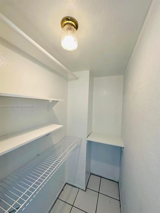 Storage space featuring white solid shelving, white wire shelving, tile flooring, and a flush-mount brass-finish light fixture with textured glass