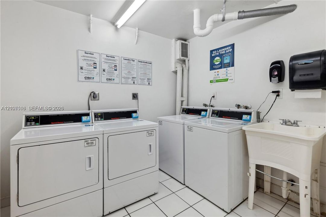 Laundry facility featuring dual white coin-operated washers and dryers, a utility sink with a dual-handle faucet, and white ceramic tile flooring
