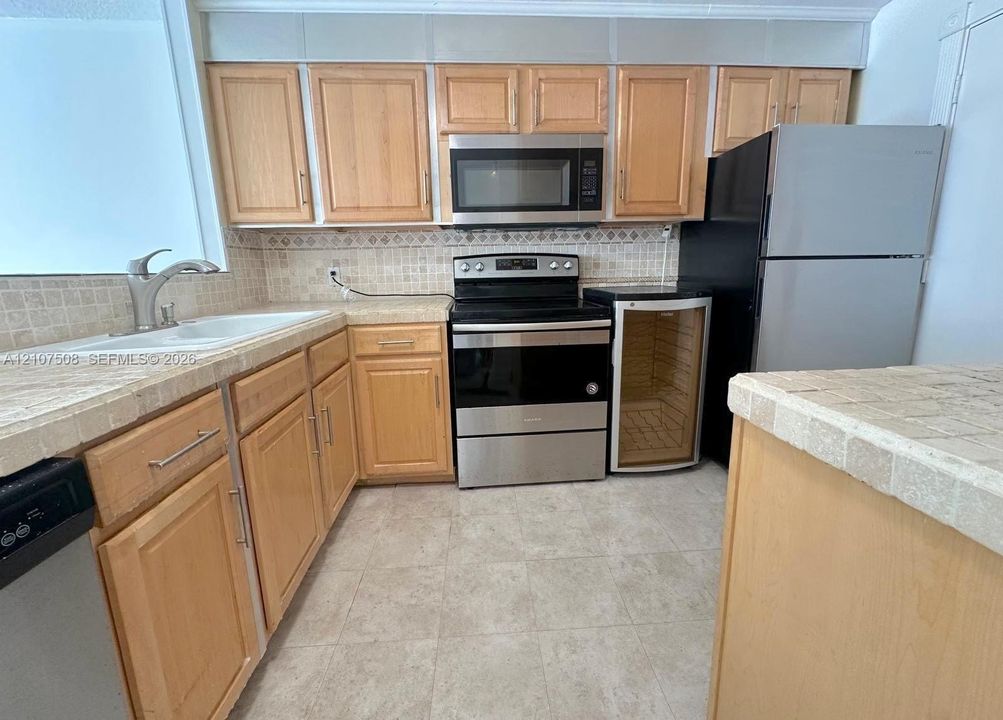 Kitchen featuring wood cabinetry, stone-finish tile countertops, mosaic tile backsplash, stainless steel appliances, and integrated wine cooler