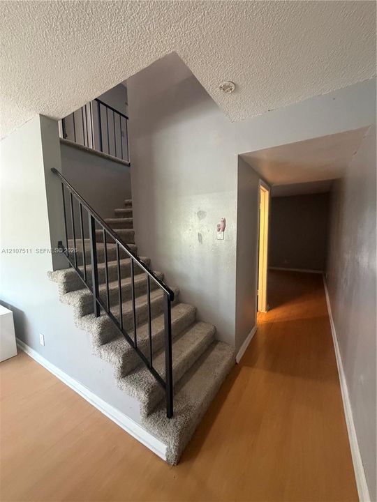 Staircase with carpeted treads and black metal railing, featuring wood-finish flooring and neutral-toned wall paint