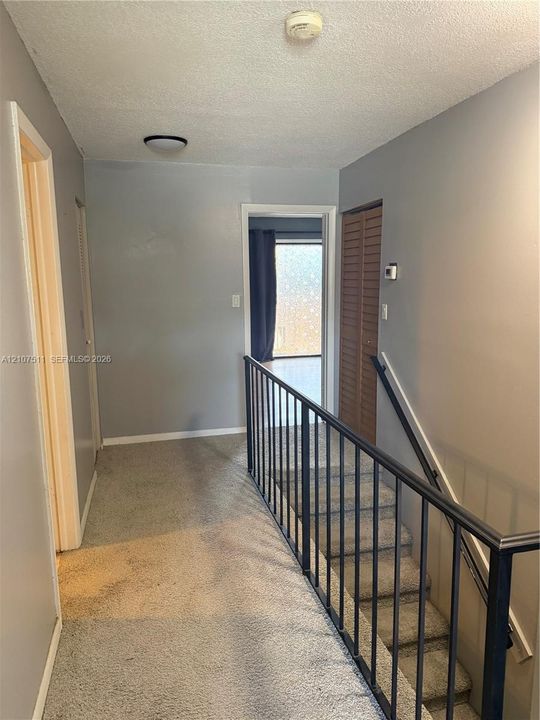 Hallway featuring neutral wall paint, carpeted flooring, and a staircase with a black metal railing