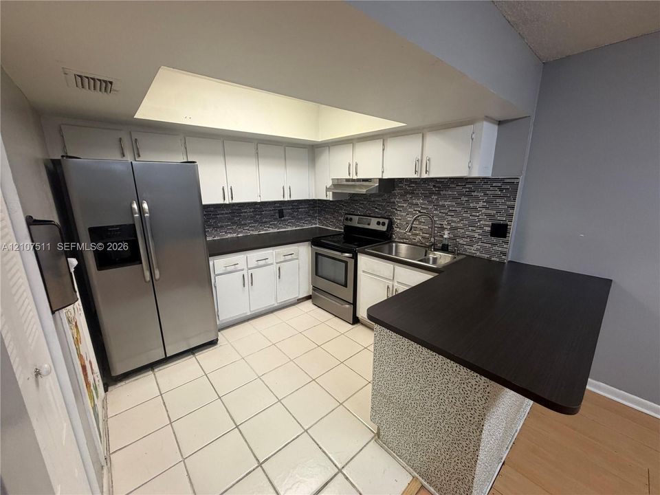 Kitchen featuring white cabinetry, stainless steel appliances, dark countertops, a stone-style tile backsplash, and a recessed ceiling light feature