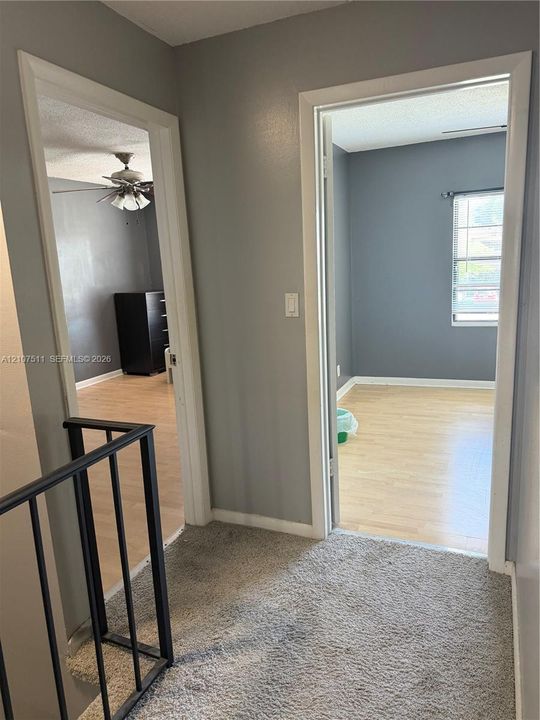 Interior hall featuring neutral-toned wall paint, wood-finish flooring in secondary rooms, carpeted hallway floor, and metal railing with vertical balusters
