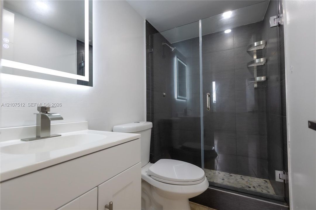 Bathroom featuring a white vanity cabinet with an integrated countertop, a backlit LED vanity mirror, and a walk-in shower with dark tile surround, a glass door, and a pebble-stone shower floor