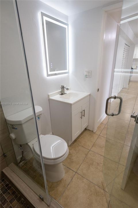 Bathroom featuring a vanity with a solid-surface countertop, an integrated sink, a frameless glass shower enclosure, a LED-backlit mirror, and tile flooring