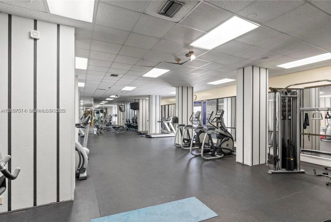 Fitness center featuring black-and-white vertical wall paneling, a suspended drop ceiling with recessed lighting, integrated wall mirrors, and rubberized athletic flooring