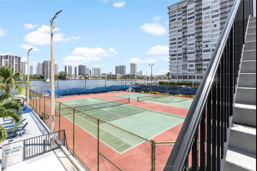 Tennis courts with chain-link fencing, paved surfaces, and net systems positioned near the water