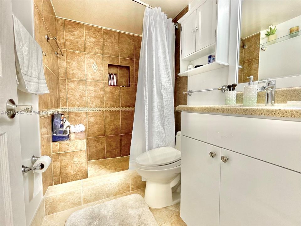 Bathroom featuring large-format tan wall tiles, built-in tiled shower bench with inset shelving, white vanity cabinetry with integrated countertop, and mounted medicine cabinet