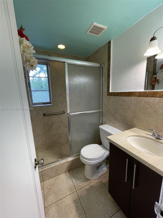 Bathroom featuring large-format stone-finish floor and wall tiles, dark wood-finish vanity with an integrated countertop, sliding glass shower enclosure, and a window with textured privacy glass