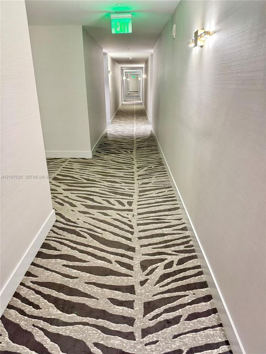 Hallway featuring textured wall coverings, patterned carpet flooring, and wall-mounted light fixtures