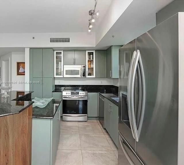 Kitchen featuring flat-panel cabinetry with glass-front accents, stainless steel appliances, dark stone countertops, tile flooring, and track lighting