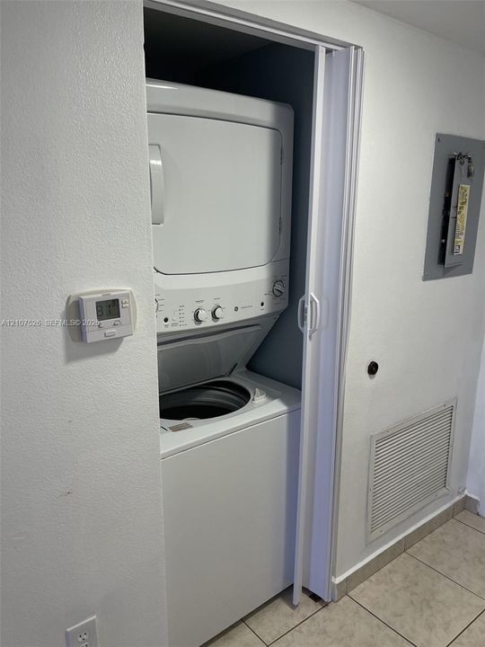 Stacked laundry unit set within a closet, featuring bi-fold doors, wall-mounted digital thermostat, and neutral tile flooring