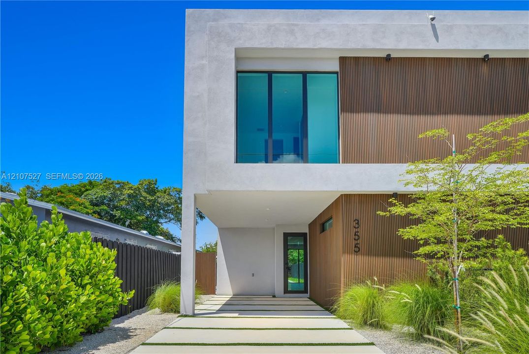 Contemporary architectural exterior featuring smooth stucco finish, vertical wood-panel siding, expansive floor-to-ceiling glass windows, and a structured concrete paver driveway with integrated ground cover