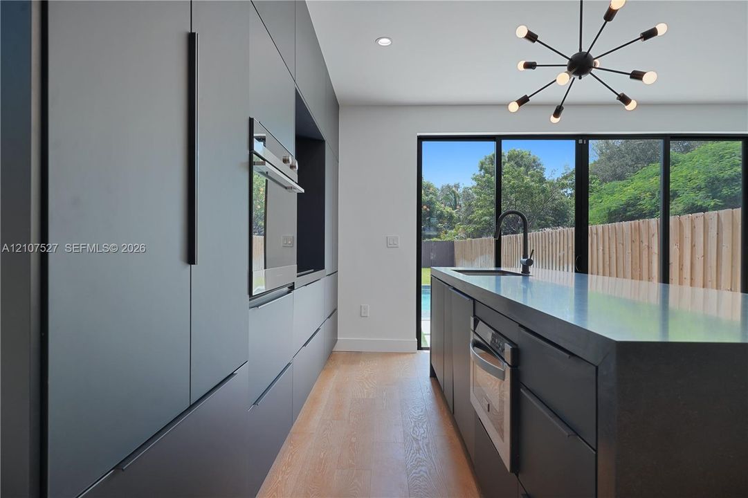 Modern kitchen featuring charcoal slab-front cabinetry, an expansive island with a matte black faucet, integrated stainless steel appliances, wood-finish flooring, and multi-panel glass doors