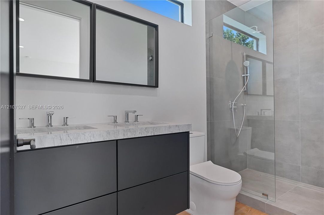 Floating dual-sink vanity with a stone countertop, framed mirrors, glass-enclosed shower with large-format tile, and rectangular transom windows