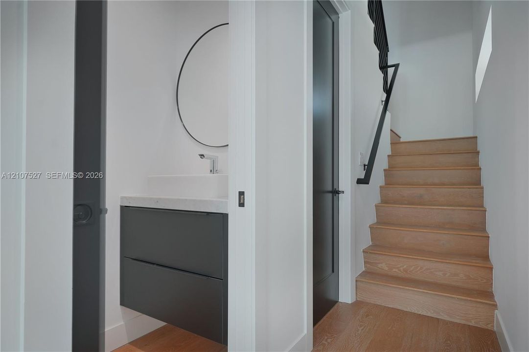 Floating vanity with stone countertop, circular wall-mounted mirror, wood-finish staircase with metal handrail, and solid-core doors