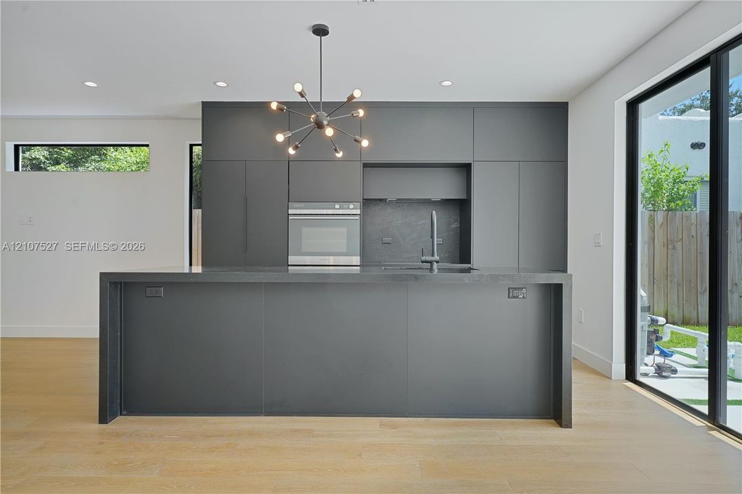 Kitchen featuring matte charcoal flat-panel cabinetry, a central island with a waterfall countertop, integrated stainless steel oven, multi-arm chandelier, and wood-finish flooring