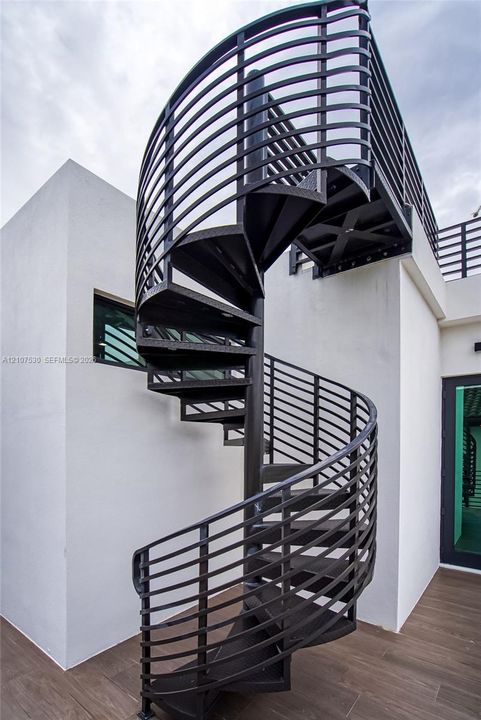 Spiral staircase featuring black metal construction with horizontal railing, smooth white stucco exterior walls, and wood-finish tile flooring