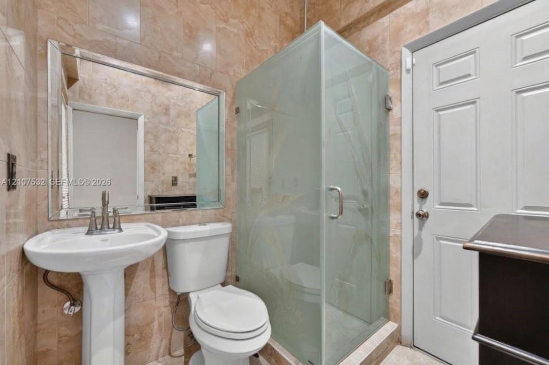 Full-height stone wall tiling, a pedestal sink with a dual-handle faucet, a framed wall mirror, a glass-enclosed shower with etched detail, and a raised-panel door