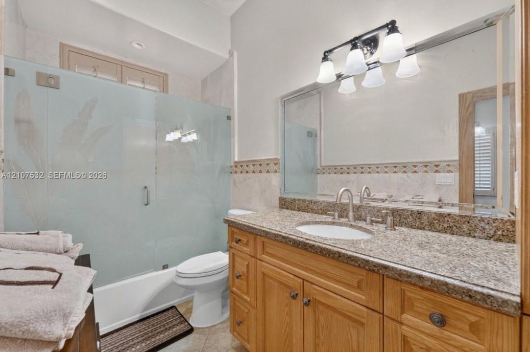 Vanity featuring wood cabinetry, stone-finish countertop, and undermount sink, complemented by a large wall-mounted mirror, four-light vanity fixture, and a tub-shower enclosure with frosted glass paneling