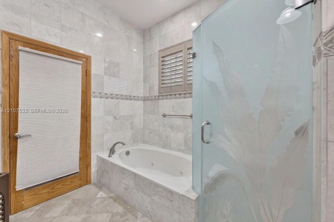 Jetted soaking tub set in a tile-surrounded deck, walk-in shower with decorative etched-glass enclosure, natural stone wall tiling with an accent border, and wood-finish door trim
