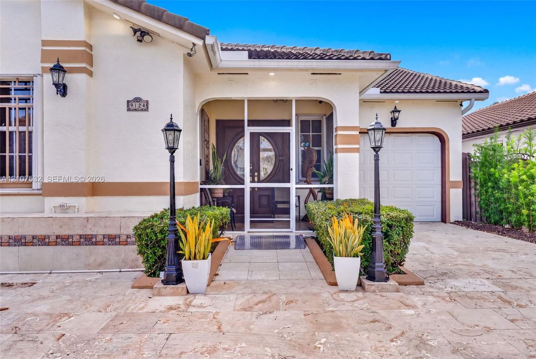 Stucco exterior featuring a tiled roof, arched entryway with a screened enclosure, ornamental iron lamp posts, and stone-finish paved driveway