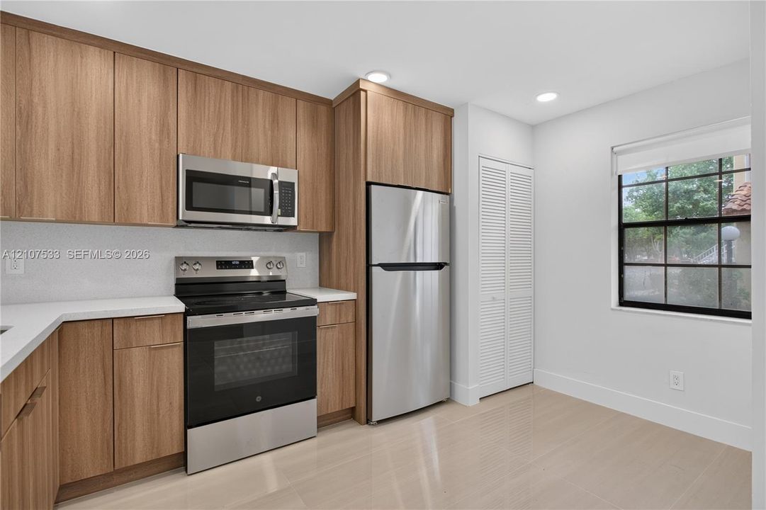 Kitchen featuring flat-panel wood-finish cabinetry, stainless steel appliances, white solid-surface countertops, tile flooring, and louvered closet doors