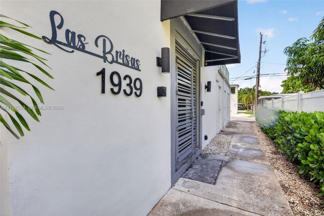 Exterior facade featuring smooth stucco finish, custom metal address signage, wall-mounted cylindrical sconces, dark metal awning, and louvered entry door