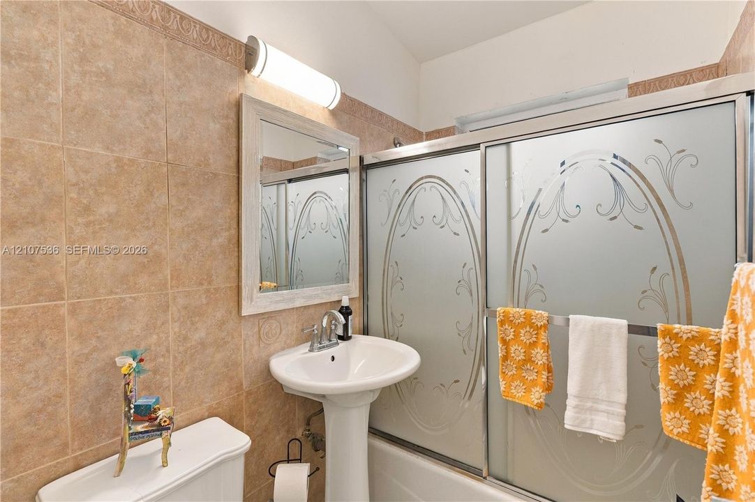 Bathroom featuring tile wall surround, pedestal sink with chrome faucet, framed wall mirror, tubular wall-mounted vanity light, and a shower enclosure with frosted glass bypass doors
