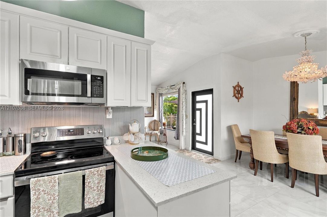 White shaker-style cabinetry, stainless steel microwave, stone-finish countertops, tile flooring, and decorative ceiling-mounted chandelier