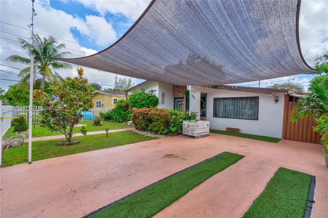 Exterior space featuring a large overhead sun shade, concrete driveway with synthetic grass inlay, white picket fencing, and mature landscaping