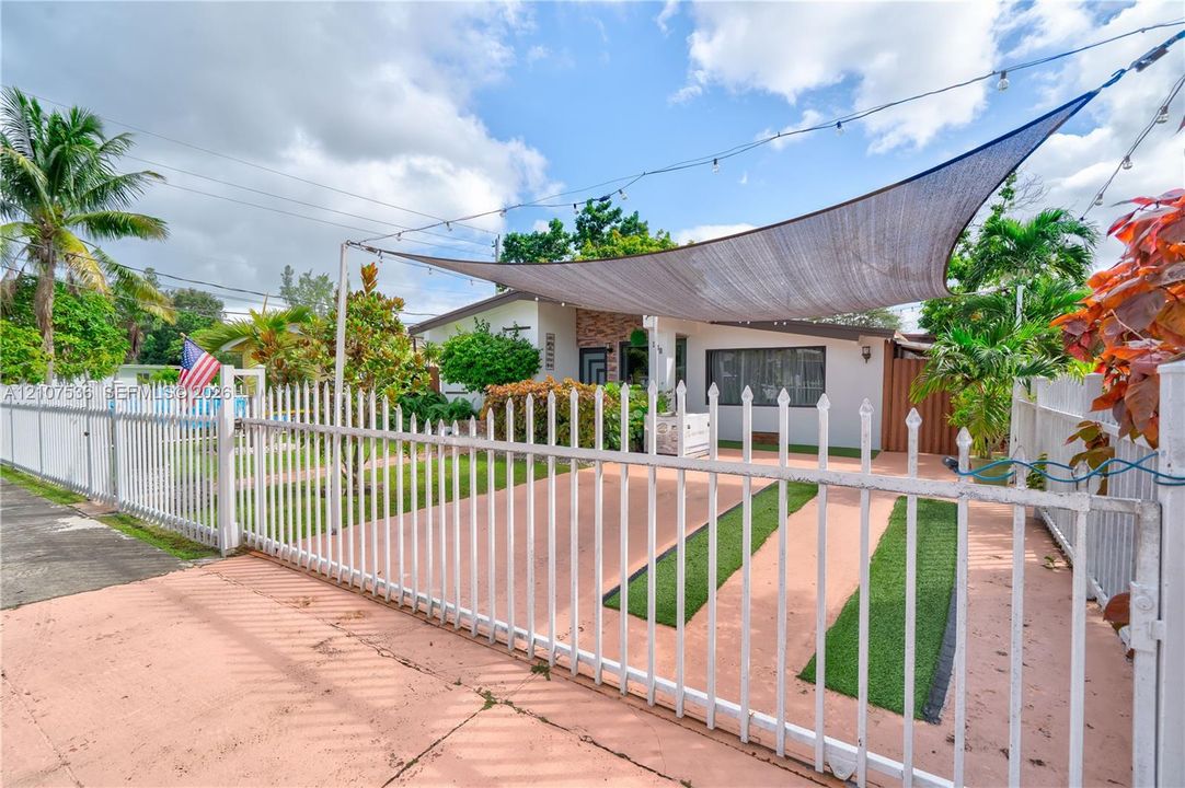 Fenced exterior featuring a concrete driveway with integrated turf strips, a shade sail installation, and white metal perimeter fencing
