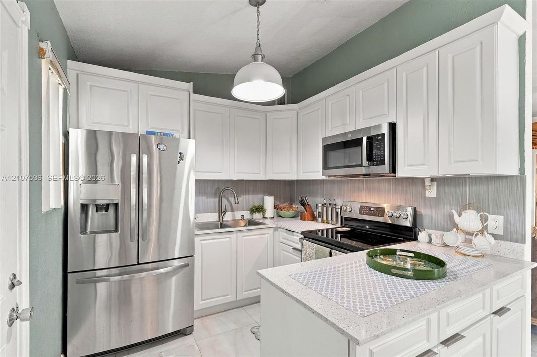 Kitchen featuring white shaker-style cabinetry, light-toned stone countertops, stainless steel appliances, vertical tile backsplash, and a pendant light fixture