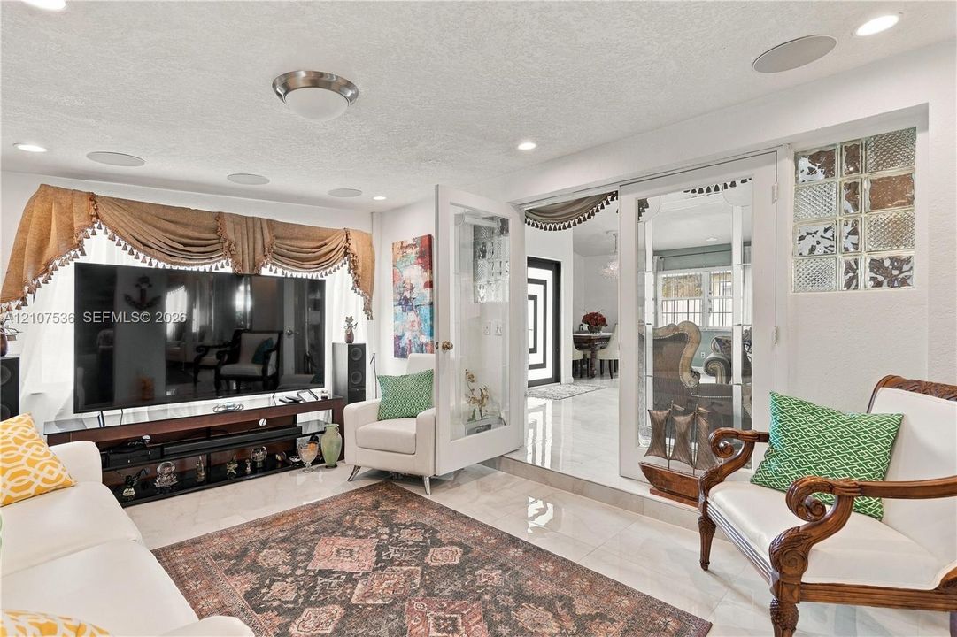 Living space featuring neutral stone-finish flooring, recessed lighting, integrated ceiling speakers, glass French doors, and decorative glass block accents