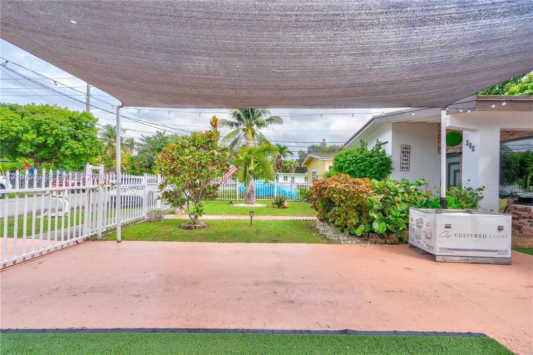 Covered concrete patio area featuring a tension shade canopy, white perimeter fencing, manicured lawn, and mature landscaping