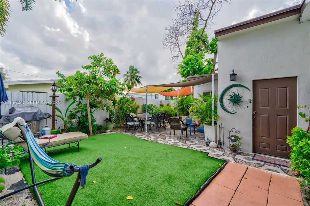Fenced backyard featuring synthetic turf, patterned concrete paver patio, attached shade canopy, and exterior wall-mounted lighting