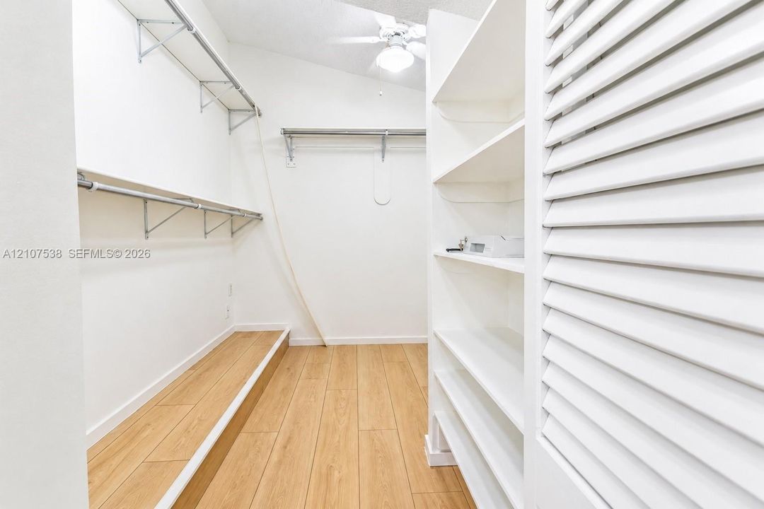 Walk-in closet featuring wood-finish flooring, built-in shelving, metal garment rods, louvered closet doors, and an overhead ceiling fan with lighting