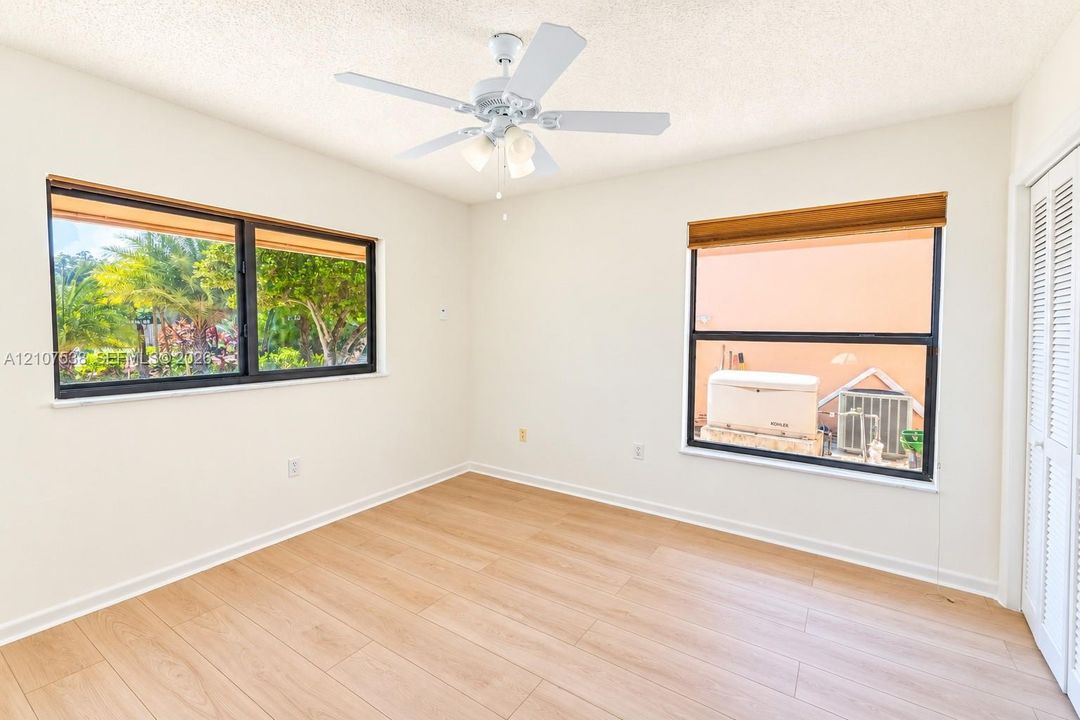 Interior space featuring light-toned wood-finish flooring, dual windows with black frames, white louvered closet doors, and a ceiling fan with integrated lighting