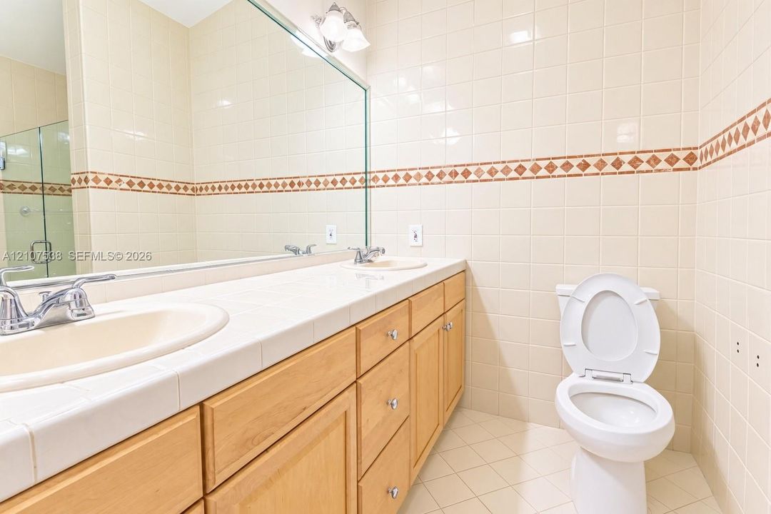 Bathroom featuring a dual-sink vanity with a tile countertop, light wood-finish cabinetry, a full-width wall mirror, and floor-to-ceiling tiled walls with decorative mosaic accents