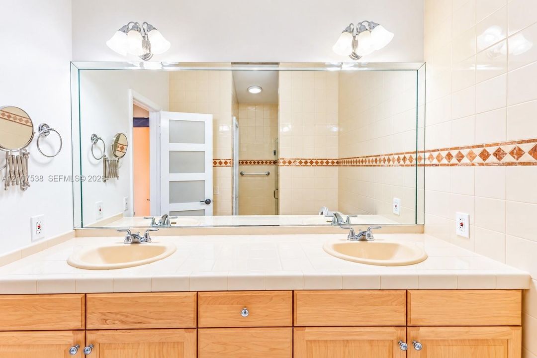 Dual-sink vanity with a tile countertop, large wall-mounted mirror, light wood cabinetry, and decorative tile backsplash