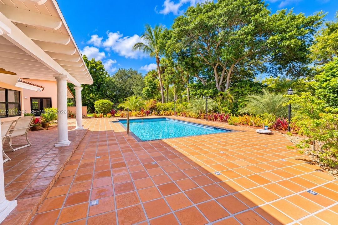 Covered lanai with white structural columns, expansive terracotta-tiled patio, rectangular swimming pool, and lush perimeter landscaping featuring mature trees and assorted tropical vegetation