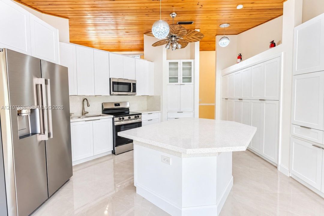 Kitchen featuring a vaulted wood-plank ceiling, white shaker-style cabinetry, a central hexagonal island with a quartz countertop, stainless steel appliances, and large-format tile flooring
